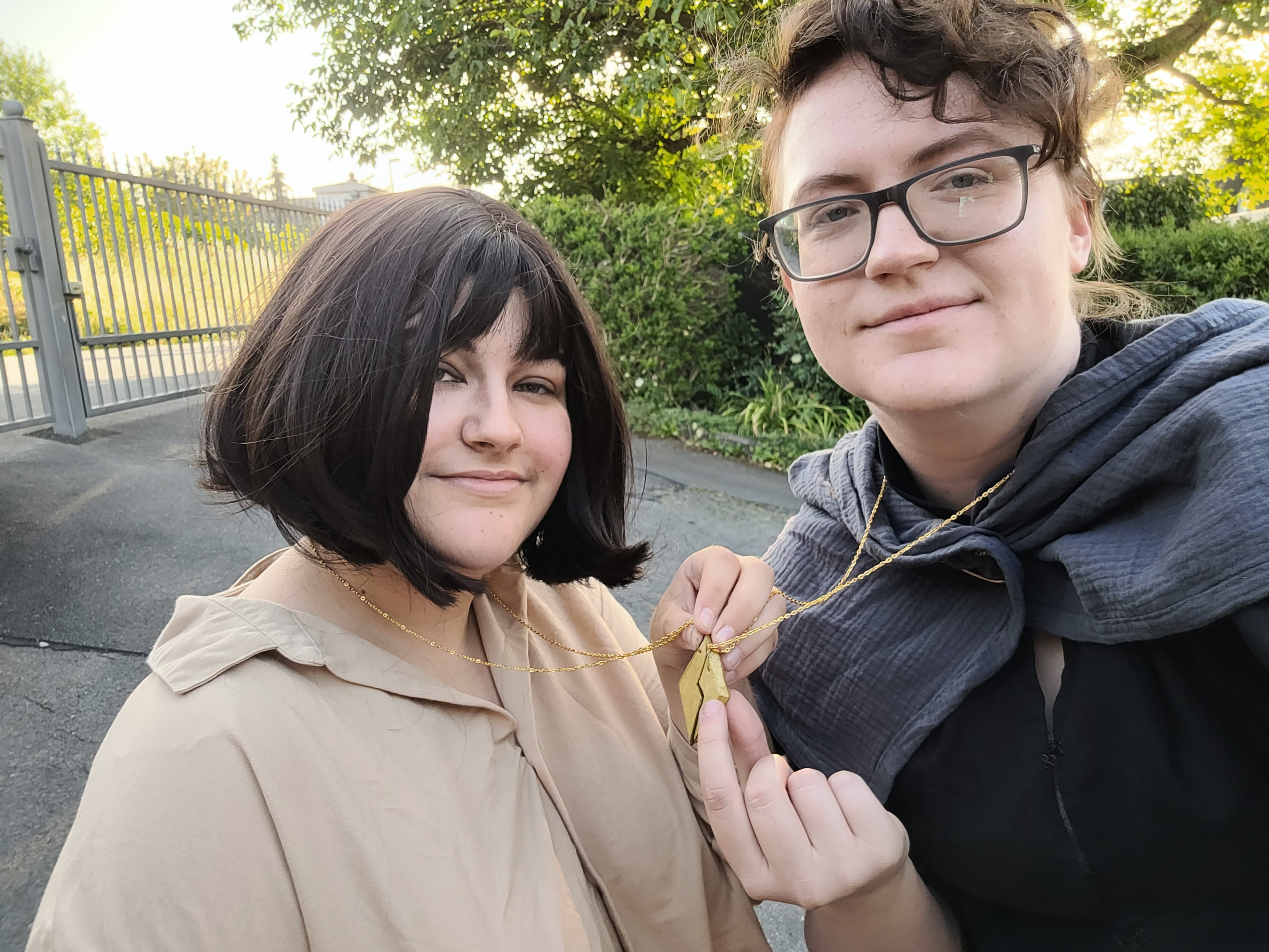 Selfie of two people cosplaying Palamedes and Camilla from the Locked Tomb. Palamedes has sort brown hair, glasses and is wearinf a grey cloak with a black shirt underneath. Camilla has chinlength brown hair and is wearing a tan shirt. Both of them are wearing a necklace. They are holding the pieces on their necklaces together like a best friend necklace, but instead of a heart it forms a gold diamond symbol, like you would find on a card deck.